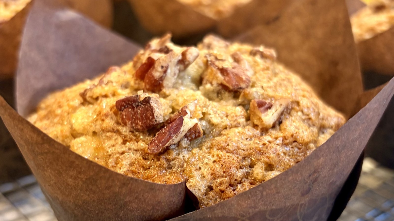 A tall Texas banana nut muffin in a brown tulip liner topped with toasted pecans, ripe bananas behind it