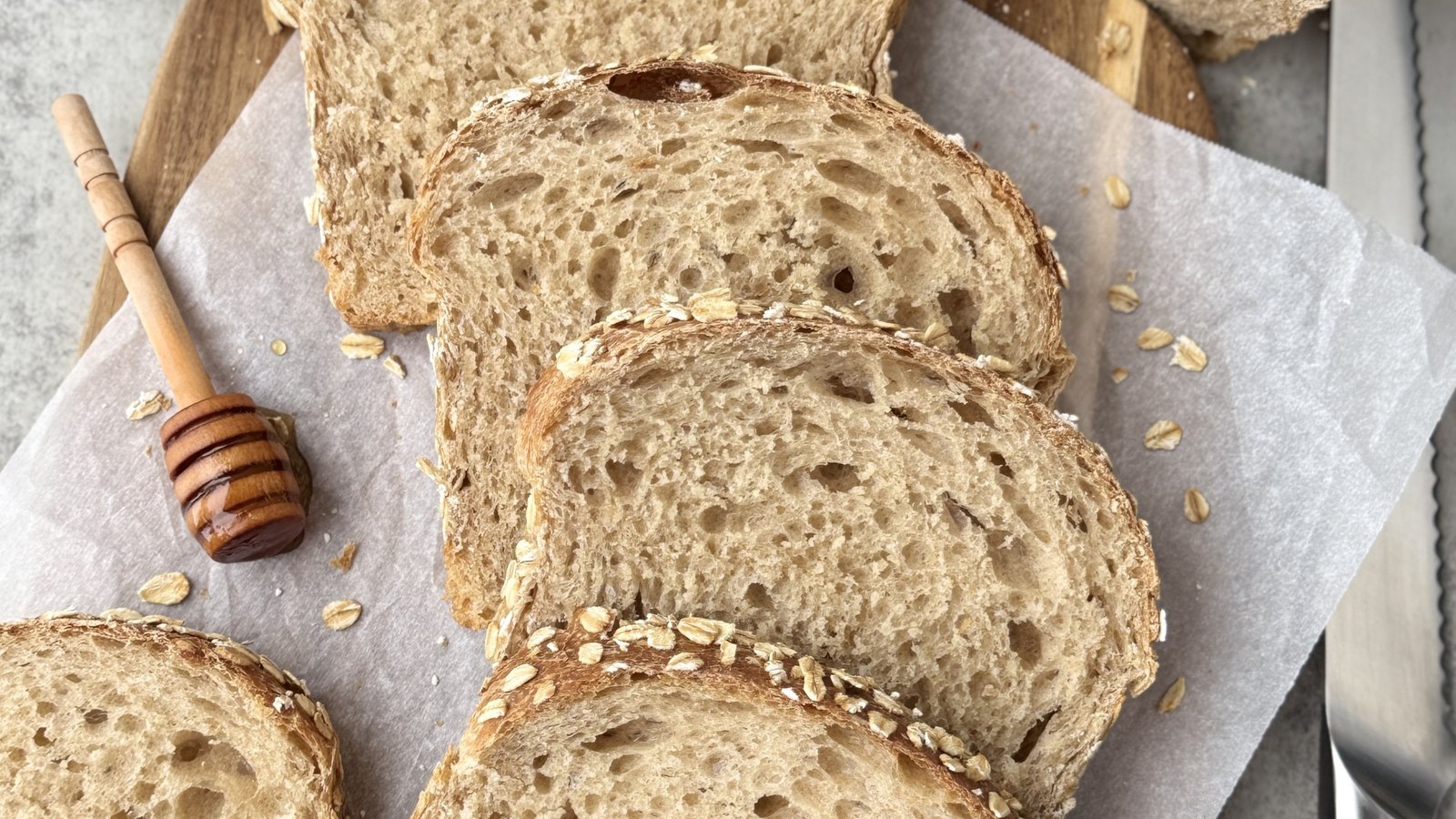 Slices of soft multigrain sourdough sandwich bread with an oat-topped crust on a wooden board beside a honey dipper